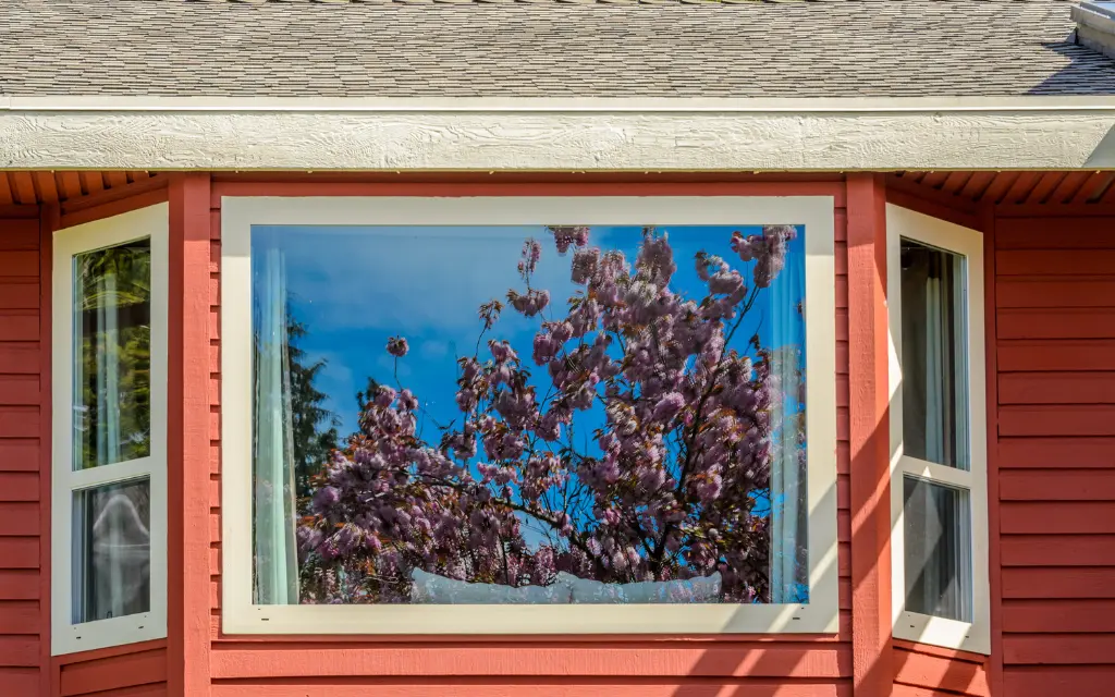 Exterior view of a bright picture bay window reflecting blooming spring cherry blossoms on a red-sided home in Toledo, Ohio.