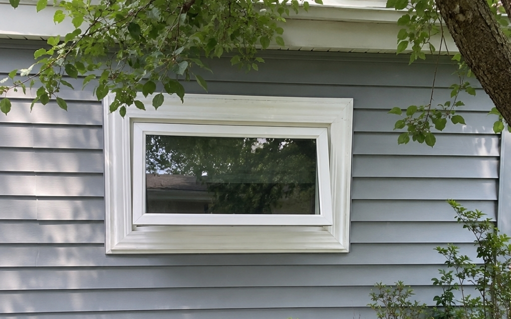White vinyl awning window installed on a gray vinyl-sided home with surrounding trees and natural shade.