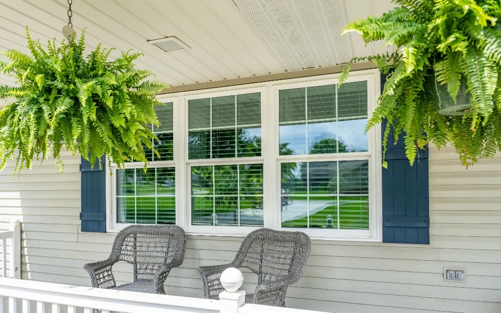 Three white-framed double-hung windows with grille inserts and navy shutters on a front porch, flanked by hanging fern baskets and wicker chairs.