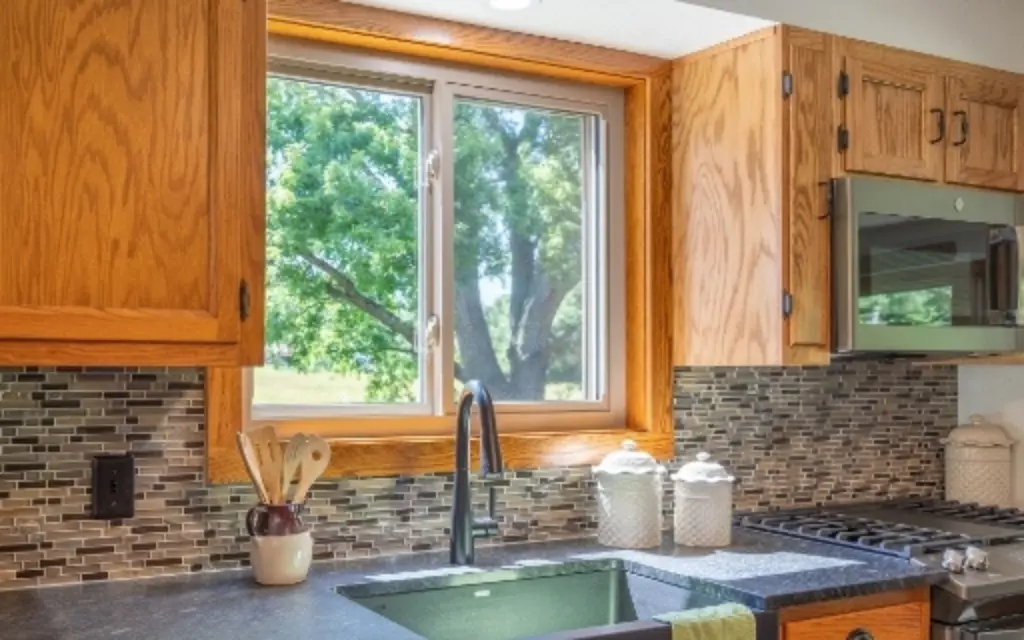 White vinyl single sliding window above a modern kitchen sink with wood cabinets and a view of mature trees.