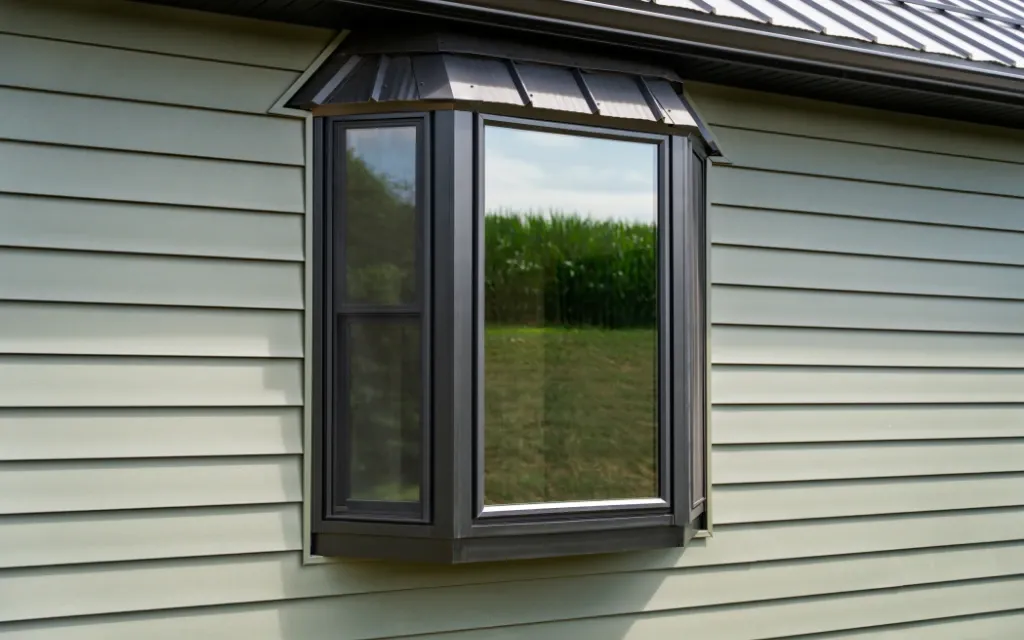 Black-framed bay window with a standing seam metal roof accent, installed on a home with light green horizontal siding, reflecting a cornfield and open sky.