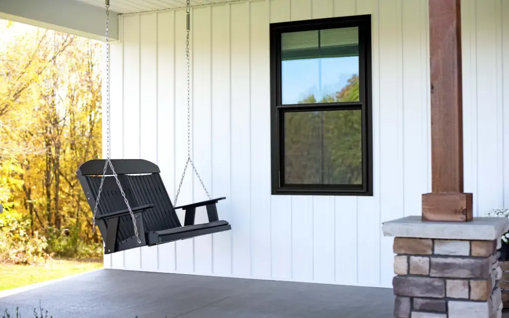 Black-framed single hung replacement window installed on a white board and batten porch wall beside a hanging porch swing, creating a clean and modern farmhouse exterior.