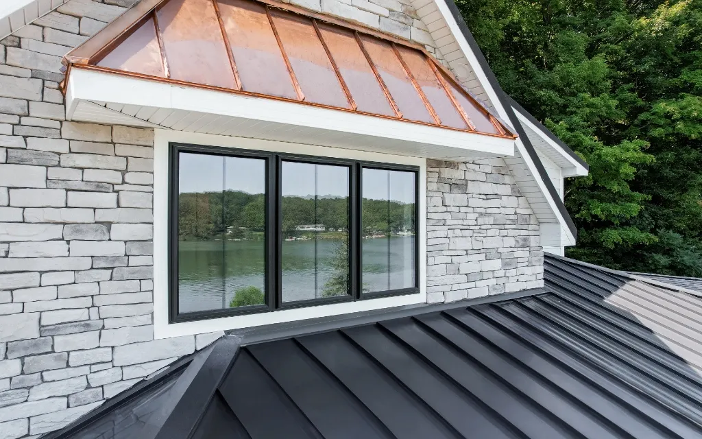 Black-framed picture window on a stone-clad dormer with a copper standing seam roof accent, reflecting a lake and tree line, with a dark metal roof in the foreground.