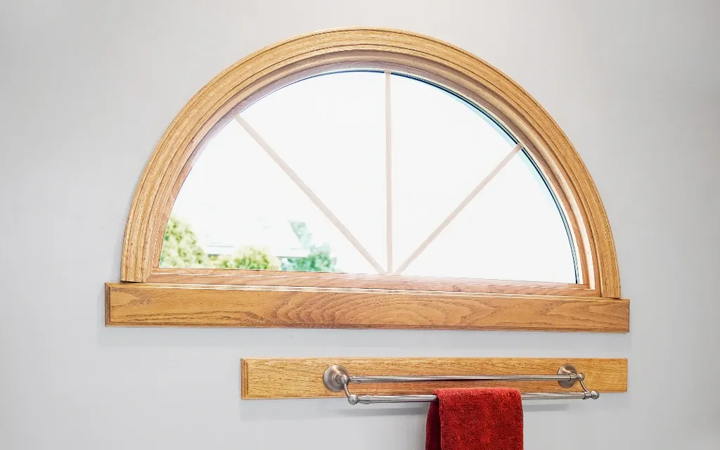 Wood-framed half-round transom window with a fan-style grille pattern above a bathroom towel bar.