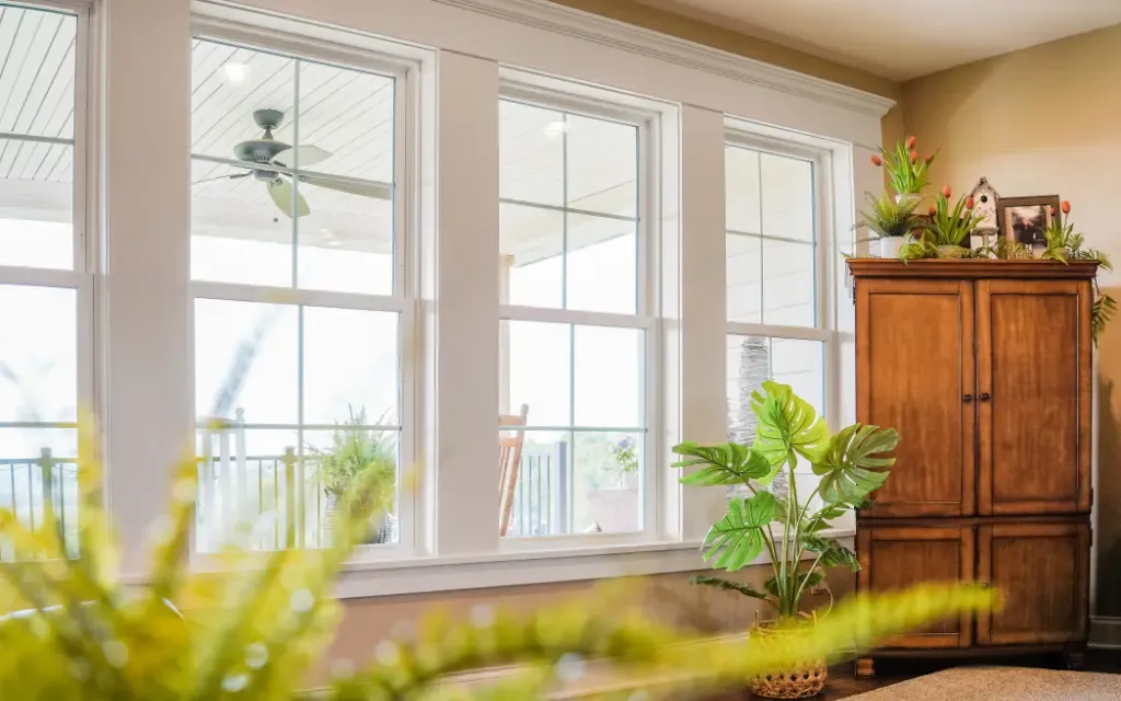 Bright living area featuring three large white double hung replacement windows overlooking a covered porch, with warm wood furniture, indoor plants, and abundant natural light.